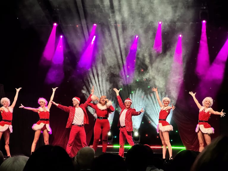 Ensemble of performers in red costumes dance under vibrant magenta lighting and vertical stage effects.