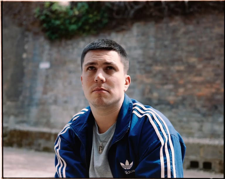 Man in blue Adidas tracksuit standing against weathered brick wall.