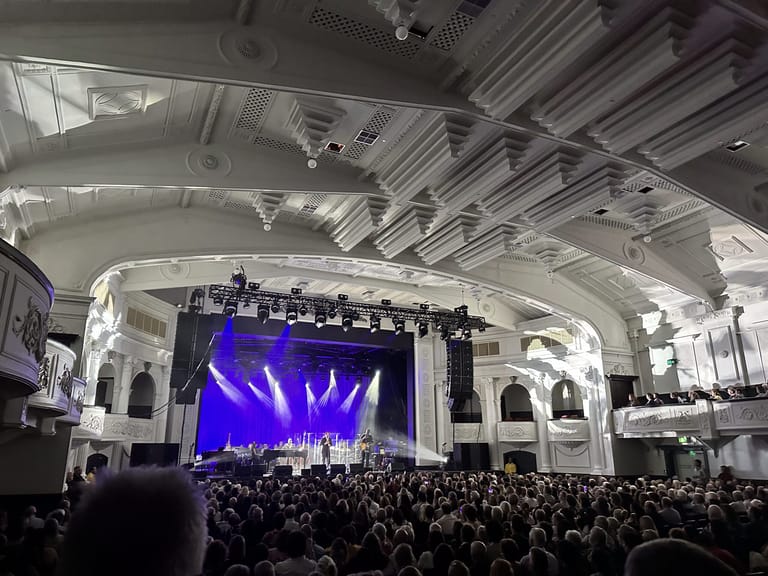 Large concert stage with purple lighting and spotlights illuminating performers, surrounded by a packed crowd in a grand theatre venue.