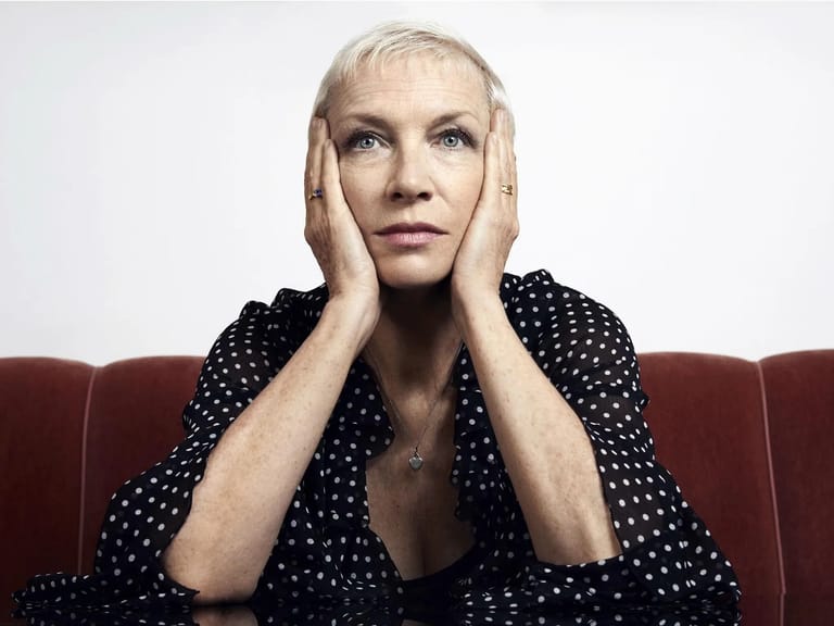 Woman with blonde hair resting her face in her hands, seated on a burgundy couch against a white background.