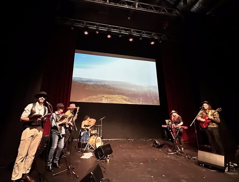 Band performing on stage with a landscape projection screen behind them in a darkened theatre.