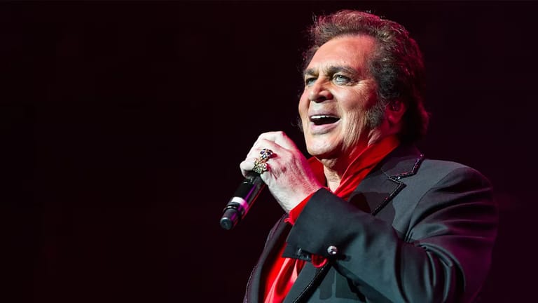 Male singer in a black jacket and red shirt holding a microphone on stage, lit by red spotlights.