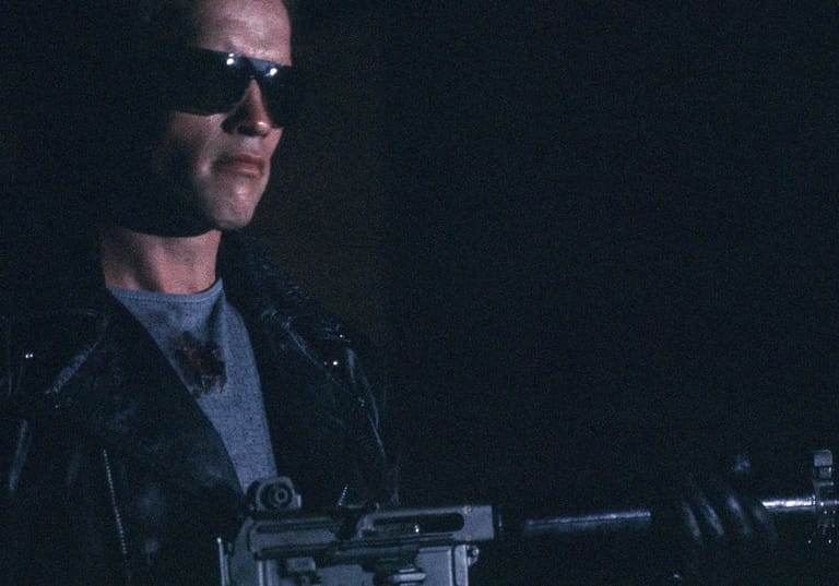 Man wearing sunglasses and leather jacket holds a futuristic weapon against a dark background.