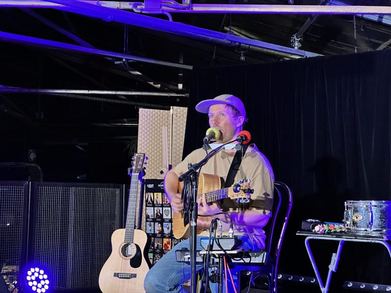 Musician performing with acoustic guitar at microphone under blue stage lighting, surrounded by loop pedals and equipment.