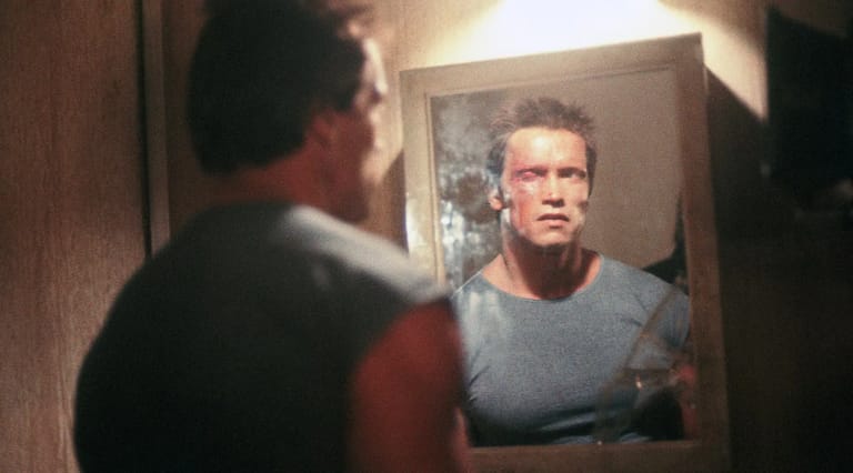 Man in grey shirt gazes at his reflection in a bathroom mirror.