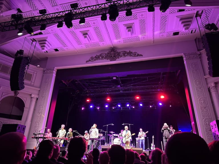 Band members perform on stage beneath purple stage lighting in a grand theatre venue, with audience members visible in the foreground.