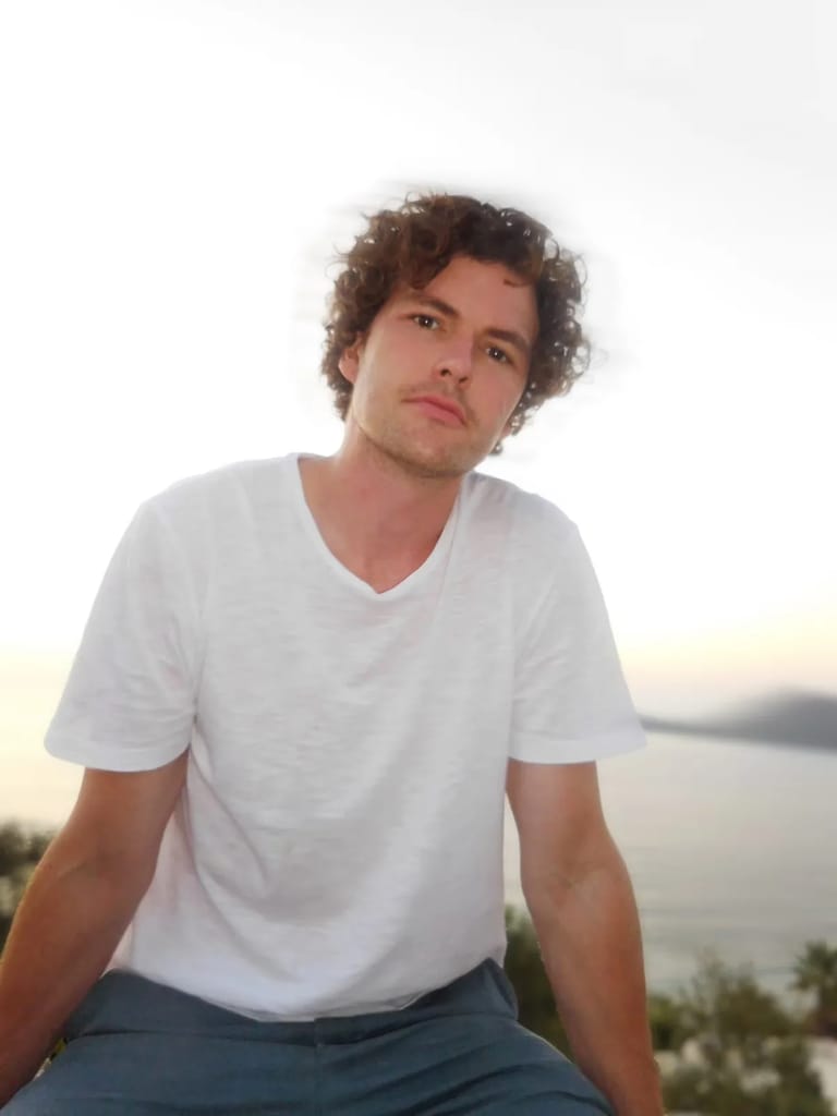 Man with curly brown hair in white t-shirt seated outdoors at dusk with water and landscape behind.