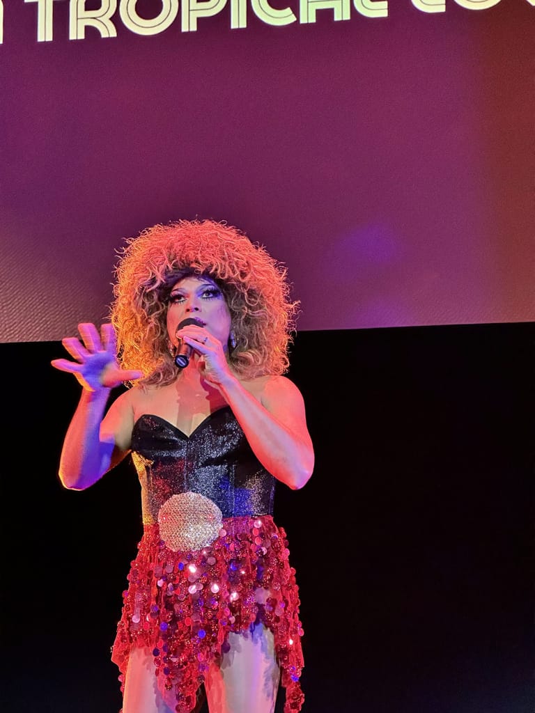 Performer in sequinned costume with voluminous orange wig sings into microphone under purple and pink stage lighting.