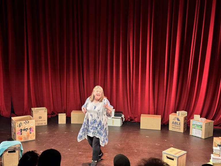 Performer in a patterned top stands centre stage surrounded by cardboard boxes against deep red theatre curtains.