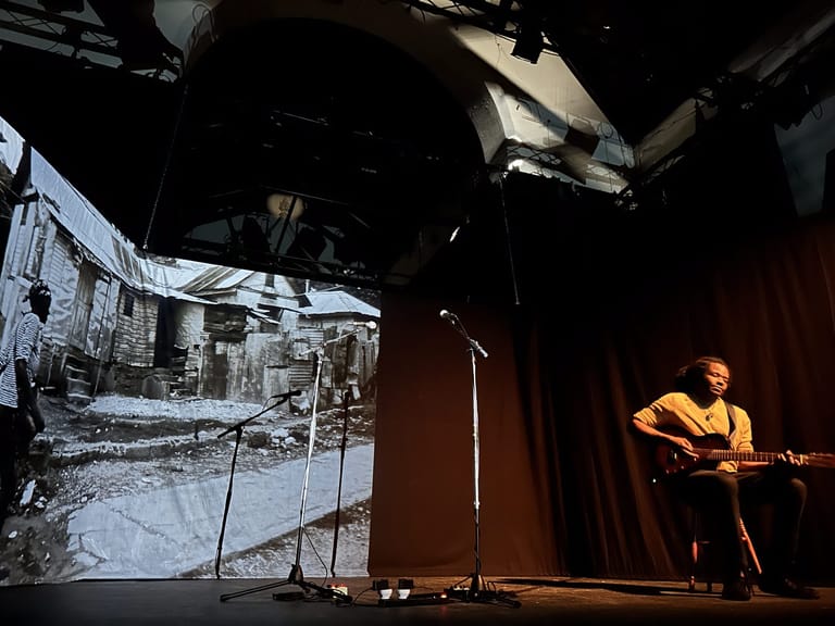 Performer with guitar on stage beside projection of a rural dwelling during a Bob Marley history presentation.