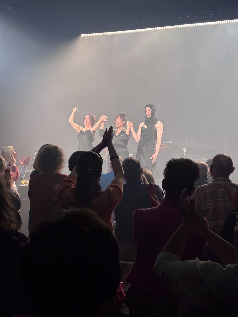 Five performers on stage engaged with a crowd during a theatre production, bathed in bright white stage lighting.