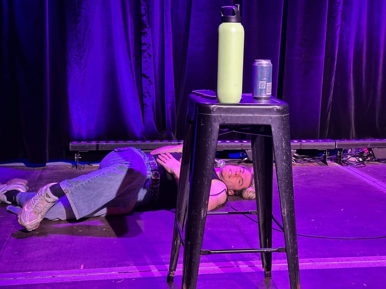 Performer lying on stage beneath a tall stool holding bottles, bathed in purple and pink stage lighting.