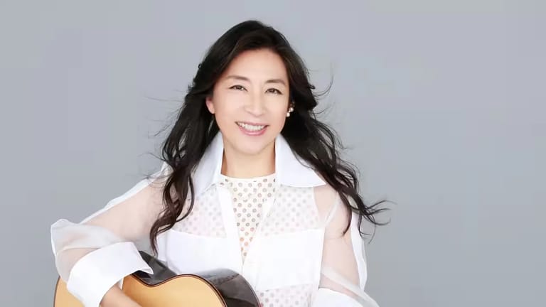 Musician holding an acoustic guitar, smiling at the camera against a grey background.