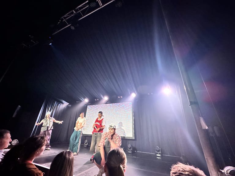 Performers on stage beneath bright spotlights during a live concert, with audience members visible in the foreground.
