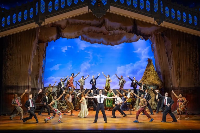 Ensemble cast in period costumes performs choreographed scene on stage with painted sky backdrop and wooden set architecture.