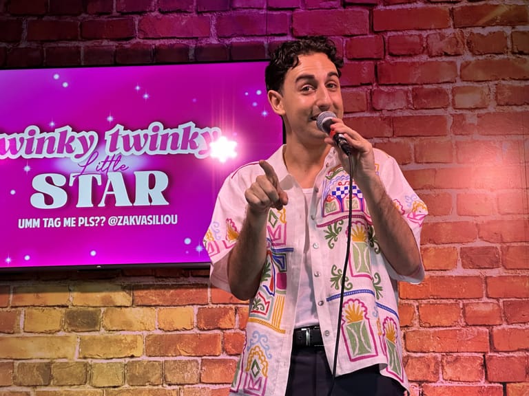 Performer in a patterned shirt speaking into a microphone against a brick wall with pink neon signage behind.