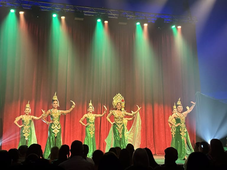 Five performers in elaborate gold and green costumes pose on stage beneath coloured spotlights and a red curtain.