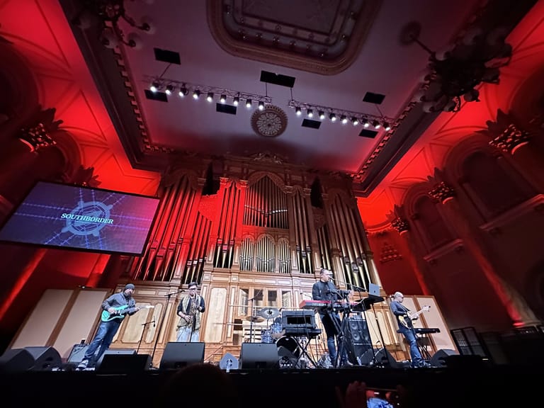 Stage set with red and purple lighting, large organ pipes, and video screens for a live music performance.