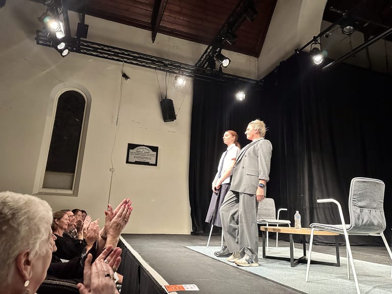 Two performers stand centre-stage during a theatrical debate, with audience members visible in the foreground.