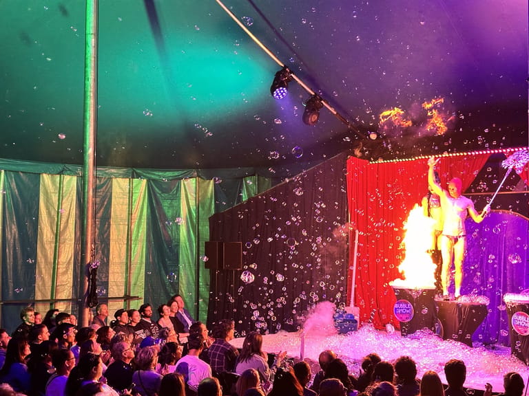 Performer creates giant bubbles on stage surrounded by colourful lighting and an engaged adult audience.