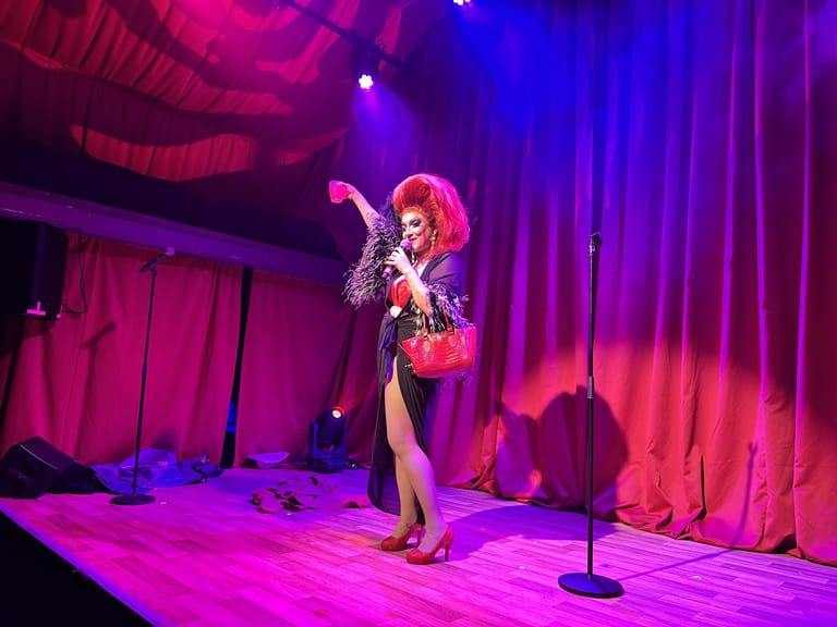 Performer with bright pink hair singing on stage beneath vibrant magenta and blue lighting.