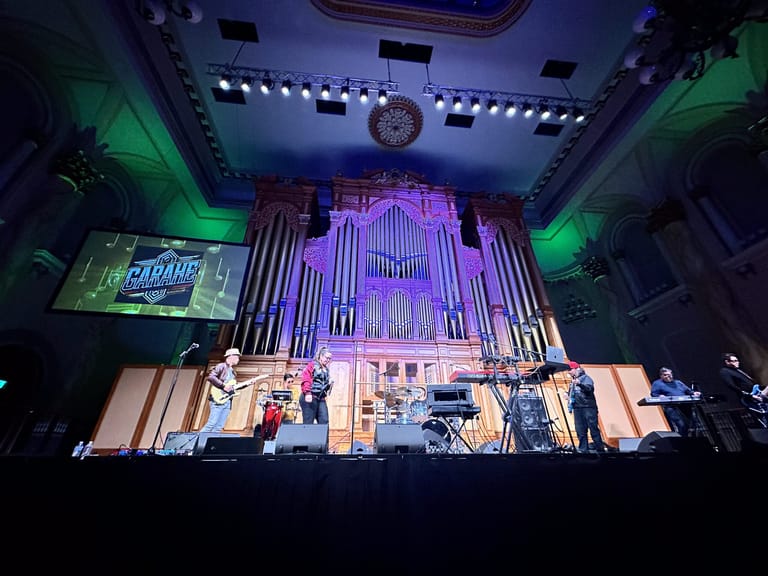 Band performing on stage beneath a large purple-lit pipe organ in a concert venue with green ambient lighting.