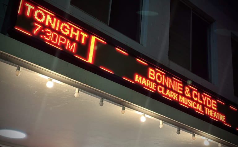 Neon marquee advertising "Bonnie & Clyde" by Marie Clark Musical Theatre, glowing red against evening darkness.