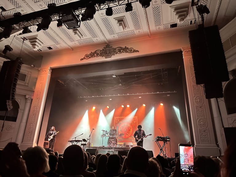Band performing on stage with orange and warm lighting, silhouetted audience members in foreground.