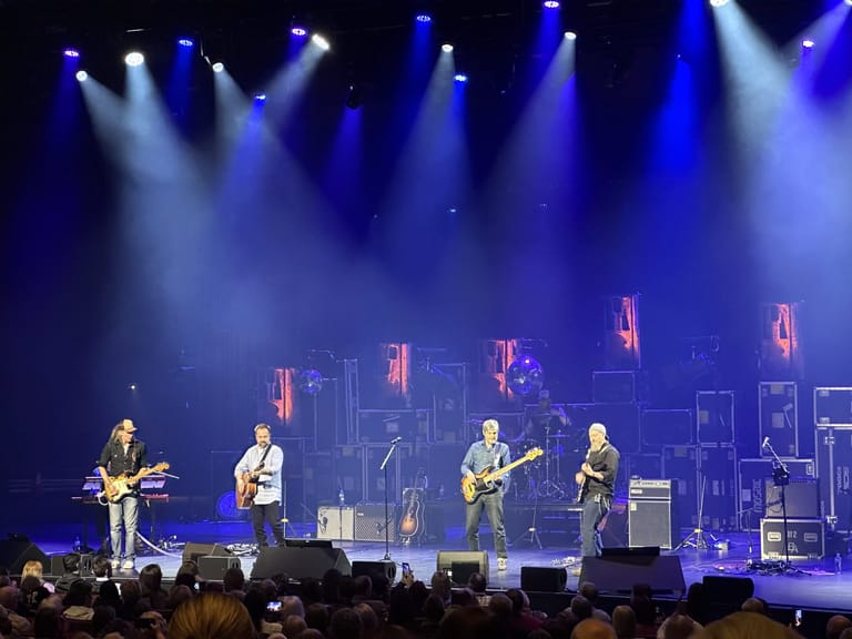 Four musicians perform with guitars on a stage bathed in blue spotlights during a concert.