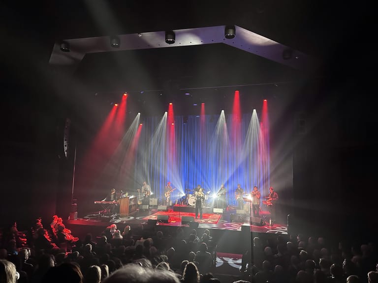 Stage bathed in red and blue lighting with band performing to a seated audience in an intimate theatre setting.