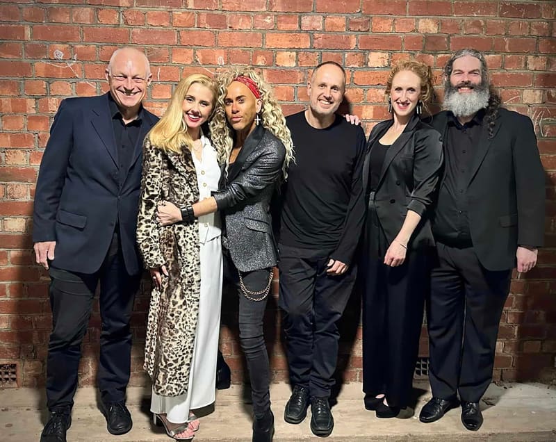 Six performers in formal black attire stand together against an exposed brick wall backdrop.