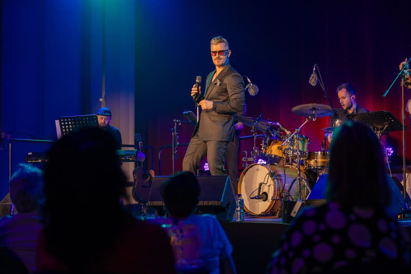 Performer in tan suit singing at microphone on stage with band and audience silhouettes in foreground.