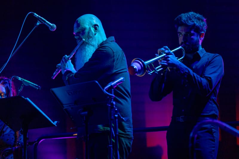 Performer at microphone with trumpet player on stage, bathed in blue and purple stage lighting.