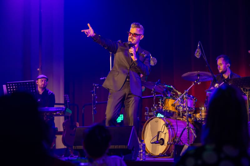 Performer in tan suit singing at microphone with band members and drummer visible under purple stage lighting.