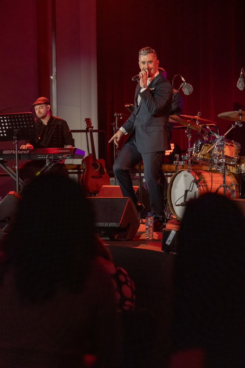 Singer in dark suit performing at microphone centre stage with band and audience silhouettes in warm red lighting.