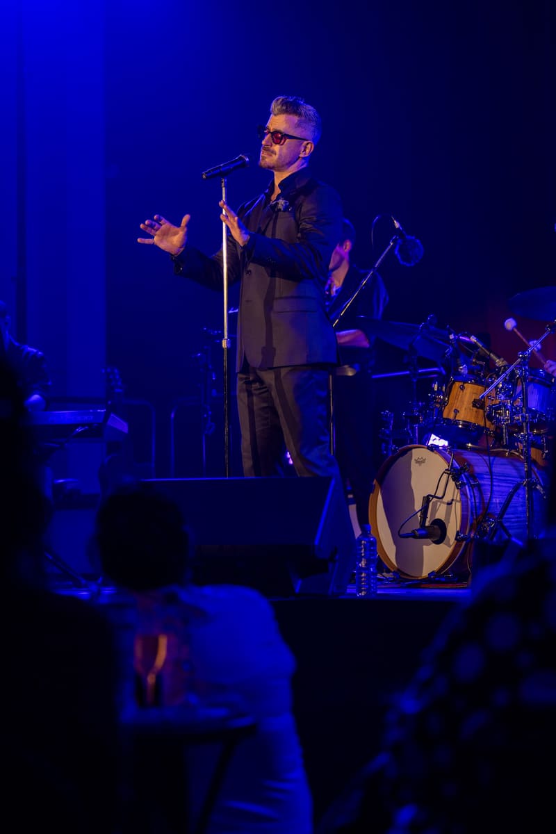 Performer in dark suit and sunglasses gestures expressively at microphone under blue stage lighting with band behind.