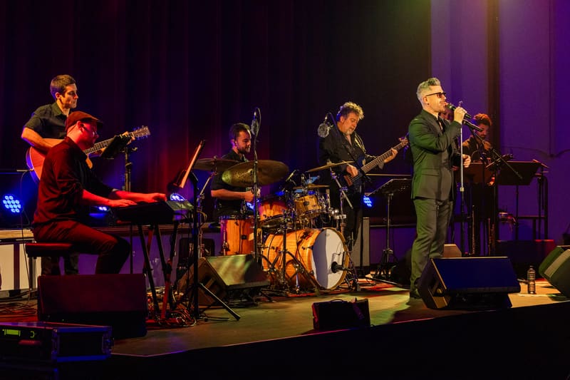 Four musicians perform on a stage lit in purple and gold, with a pianist on left, drummer centre, and vocalist in green suit on right.