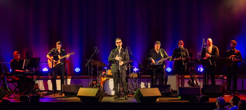 Band of musicians performing on stage with blue and purple stage lighting, centre performer in spotlight.