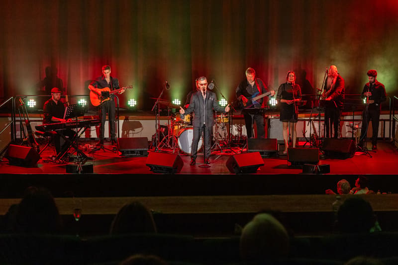 Performer in dark suit singing centre stage with band members playing instruments behind on red-lit stage.