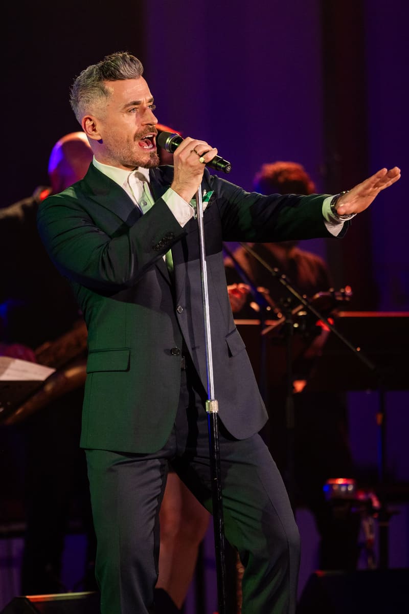 Singer in dark suit performing at microphone with arm extended, purple stage lighting and band silhouetted behind.