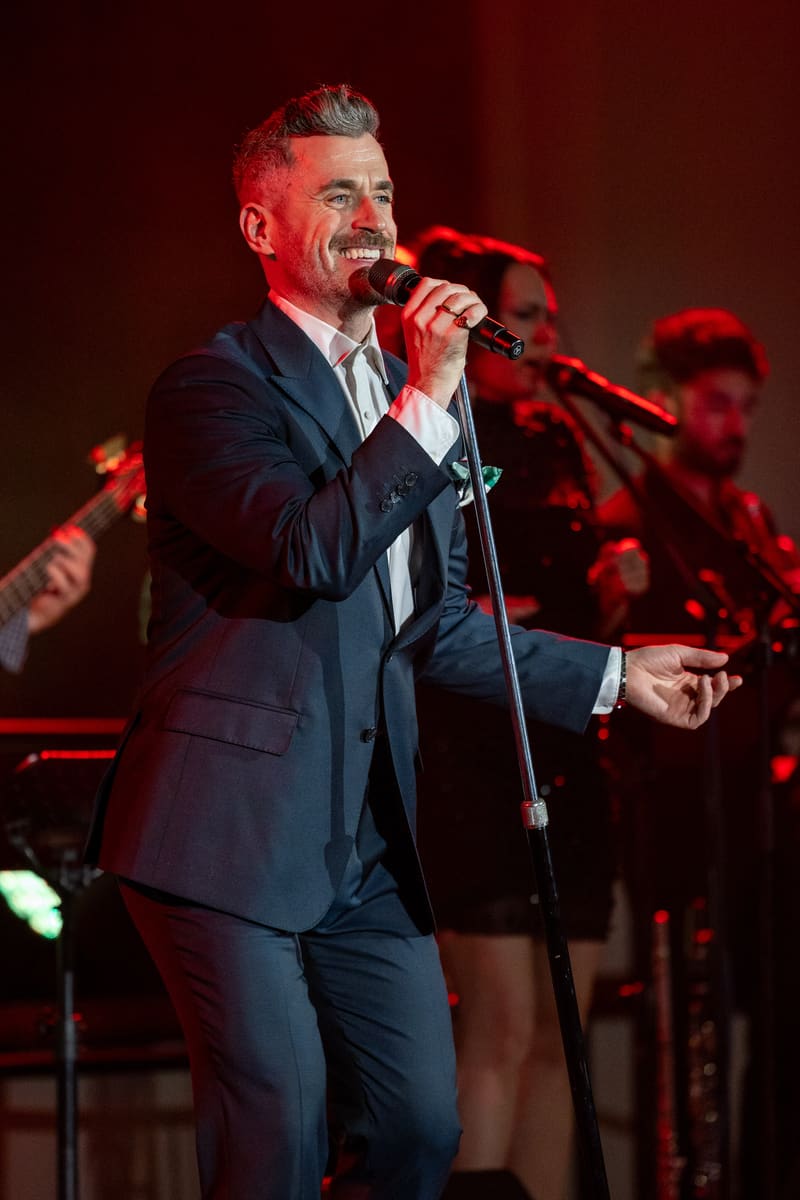 Performer in dark suit singing into microphone under red stage lighting with band members visible behind.