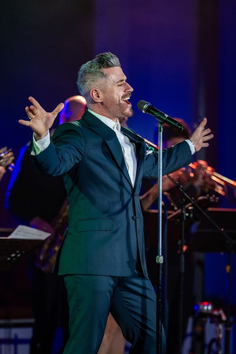Performer in dark suit singing at microphone with arms outstretched, stage lit in deep blue with band visible behind.