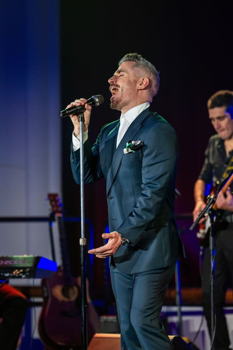 Performer in dark suit singing at microphone with band members on stage during tribute concert.