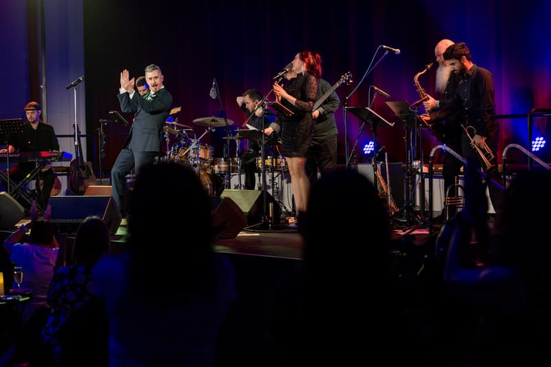 Live band performs on stage with blue lighting, audience silhouettes in foreground.