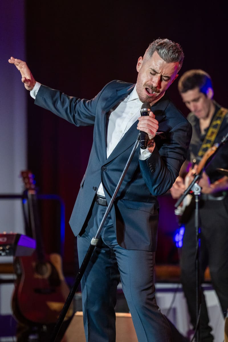 Performer in dark suit singing into microphone with arm outstretched, backed by band members on stage under purple lighting.