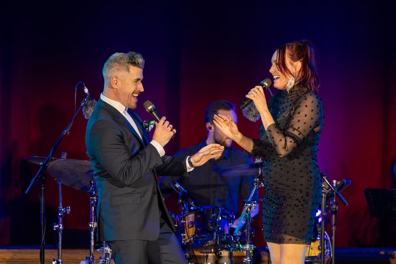 Two performers sing together on stage under red and blue stage lighting during a tribute concert.