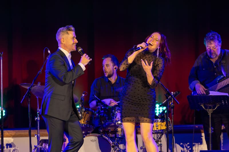 Male and female performers sing duets centre stage under red and blue lighting, backed by a live band.