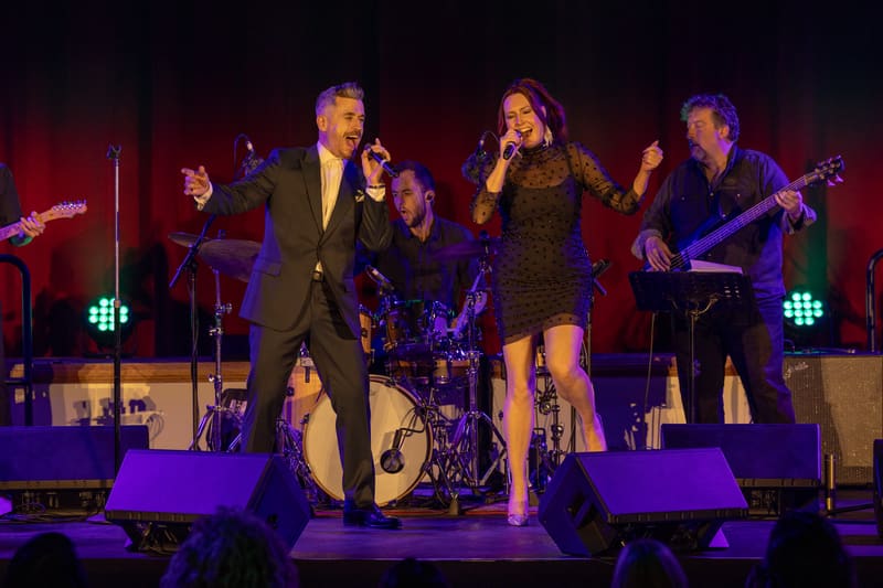 Three performers in formal attire sing and dance on a stage lit by purple spotlights during a tribute concert.