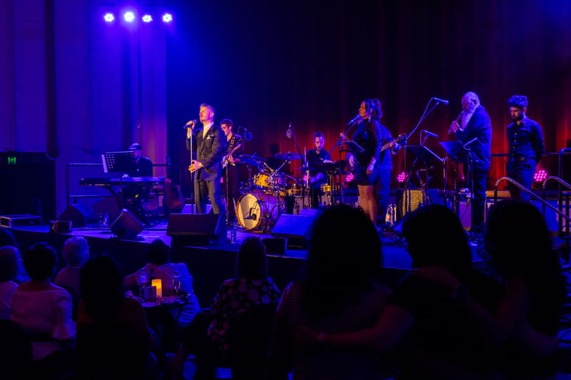 Live band performs on stage under blue spotlights with audience silhouettes in foreground.