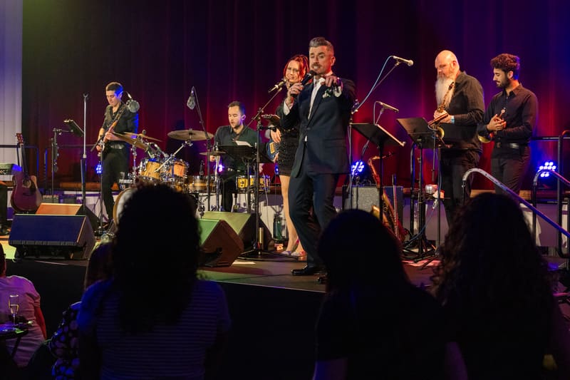 Performer in dark suit singing centre stage with live band under purple stage lighting, audience silhouettes in foreground.